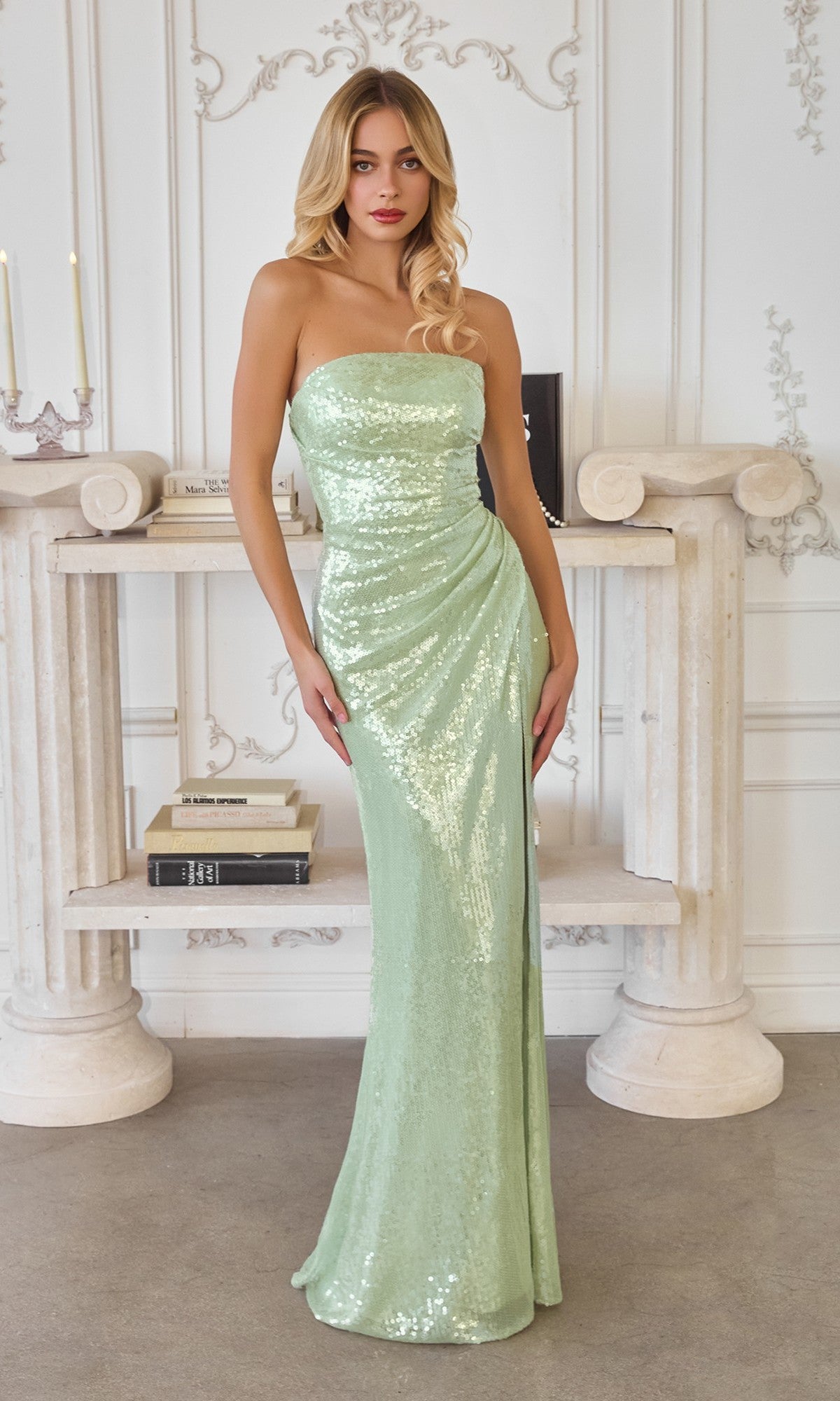 A woman wears the Amoris T724 long prom dress in sage, standing indoors before a decorative white fireplace adorned with books and candles.